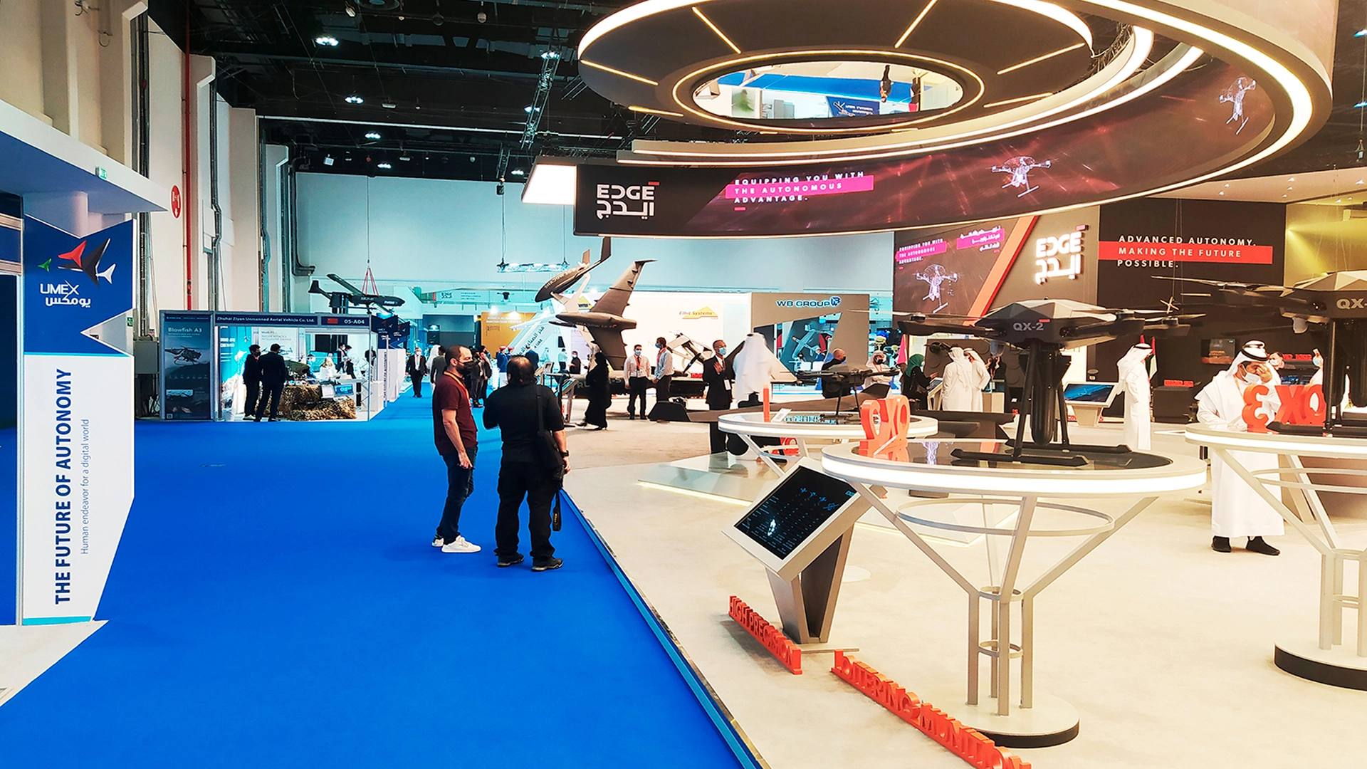 Holiday space with several people standing and several stands around. Part of the floor is blue and part is white.