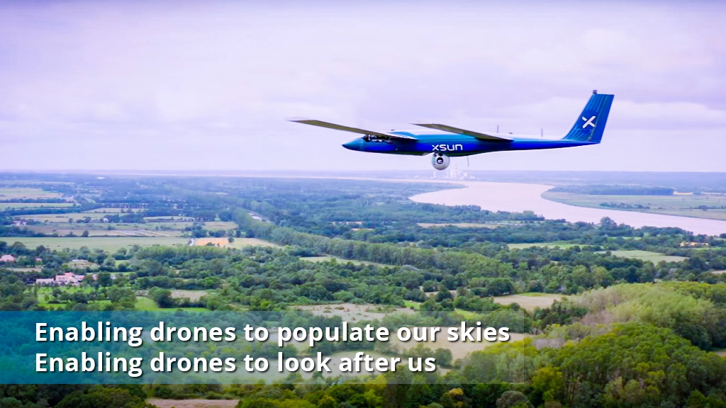 An unmanned aerial drone flies over a scenic landscape with a river and greenery, featuring the text "Enabling drones to populate our skies, enabling drones to look after us."